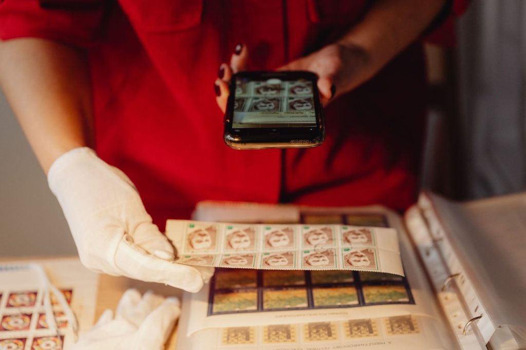 Proposta na ALMG mira regras para preservar e reutilizar conteúdos bancados pelo Estado Pessoa usando um smartphone para digitalizar uma coleção de selos com luvas